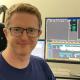 Man with blonde hair and dark rimmed glasses sits beside editing suite