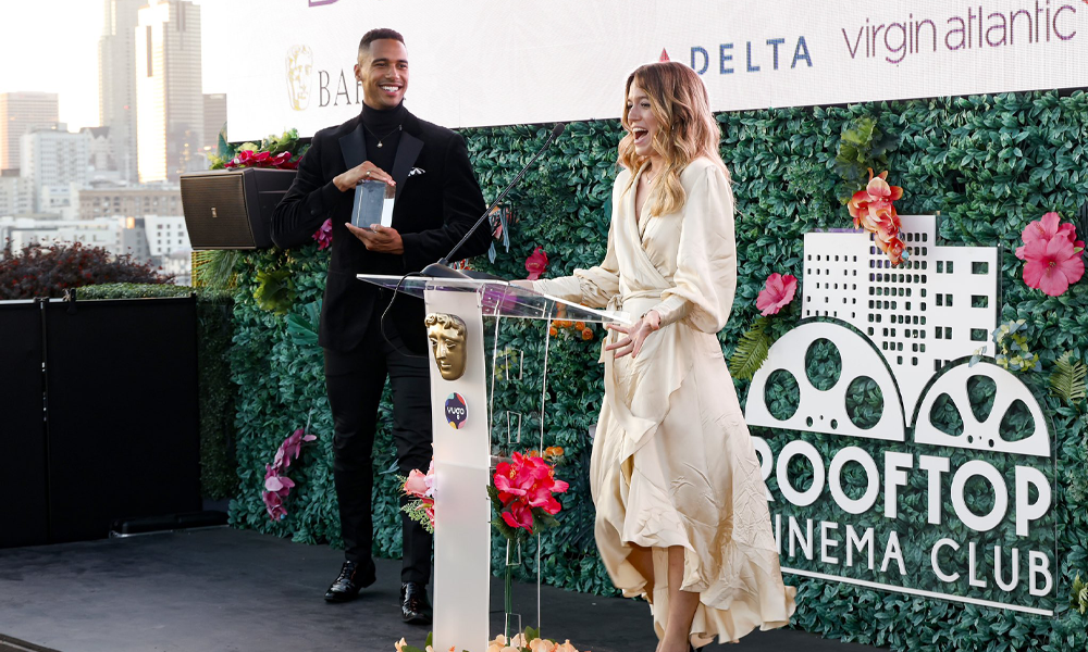 person giving speech on stage with person holding award in background