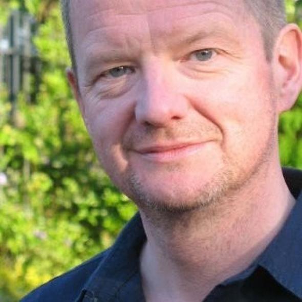 White man smiles towards camera against backdrop of trees