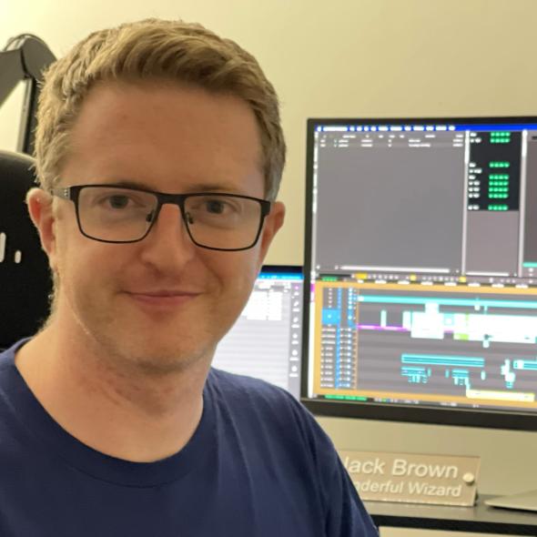 Man with blonde hair and dark rimmed glasses sits beside editing suite