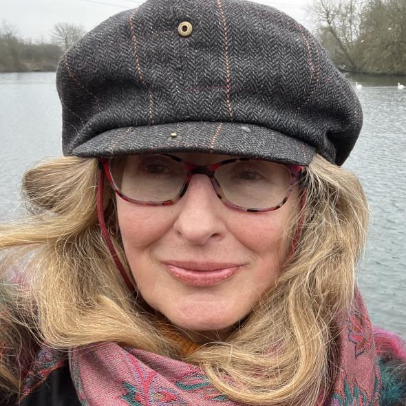 Image of woman with blonde hair wearing glasses and a large grey flat clap smiling towards camera