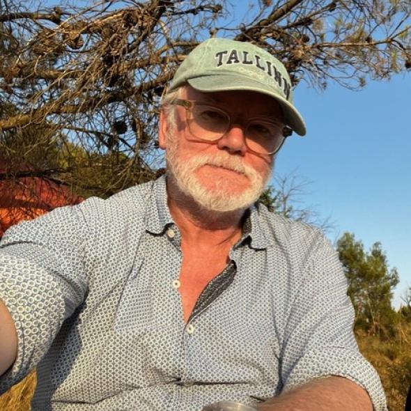 A middle-aged white man in a hat and glasses, in front of a tree