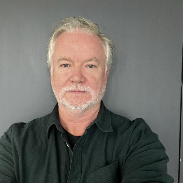 A middle-aged white man with a grey beard, standing in front of a grey background
