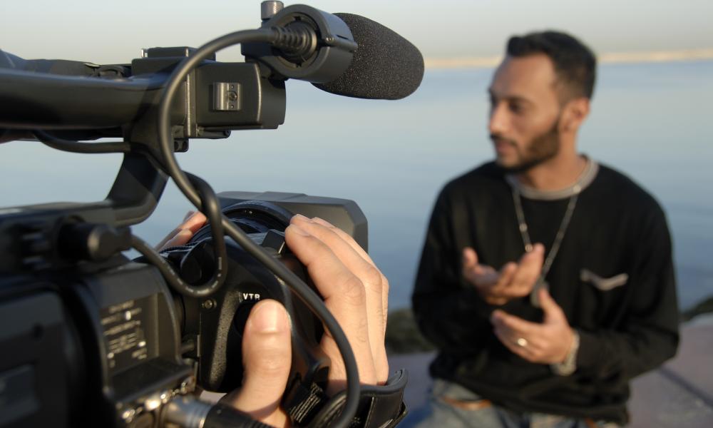 Handheld documentary camera pointing towards man speaking in background